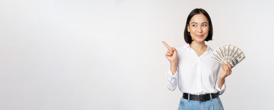 Smiling young modern asian woman, pointing at banner advertisement, holding cash money dollars, standing over white background.