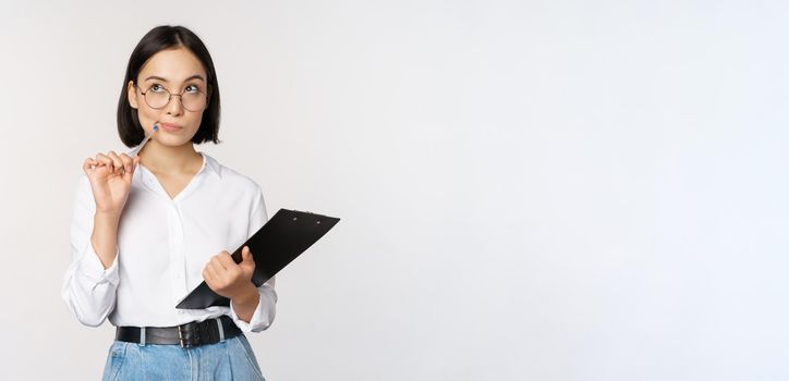 Asian girl in glasses thinks, holds pen and clipboard, writing down, making notes, standing over white background.