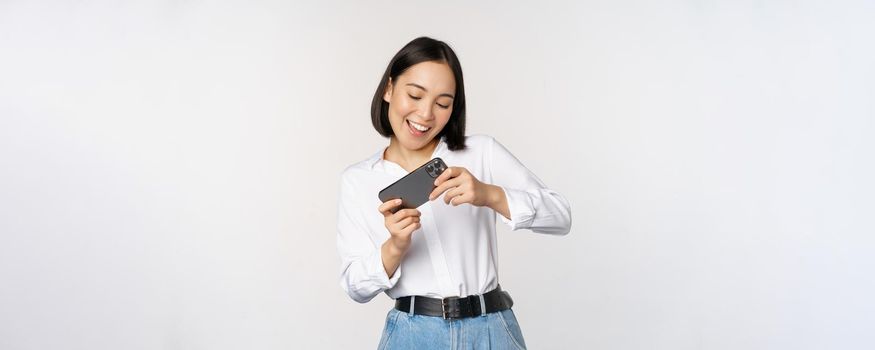 Young korean woman, asian girl playing mobile video game on smartphone, looking at horizontal phone screen, standing over white background.