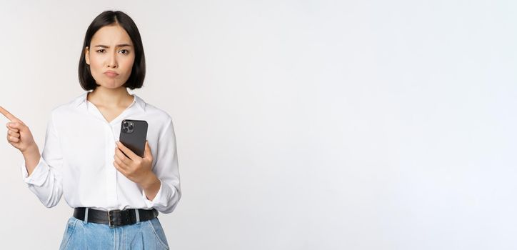 Skeptical asian modern girl, holding smartphone, pointing finger left at empty space and frowing hesitant, has doubts, standing over white background.