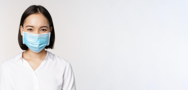 Covid and healthcare concept. Close up portrait of asian woman, office lady in face mask, smiling, using protection from coronavirus during pandemic, white background.