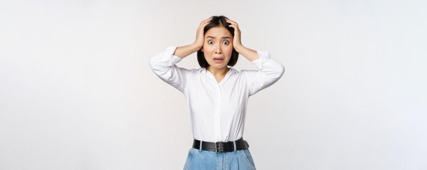 Image of shocked anxious asian woman in panic, holding hands on head and worrying, standing frustrated and scared against white background.