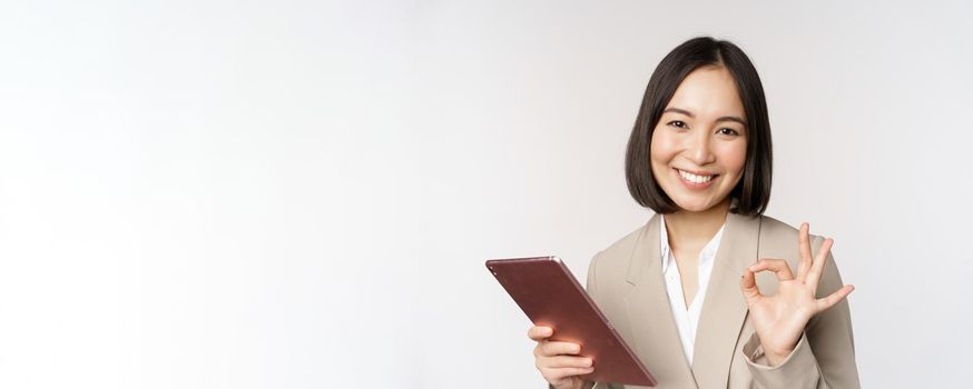 Image of successful asian businesswoman holding digital tablet, showing okay, ok sign, assuring client, standing over white background.