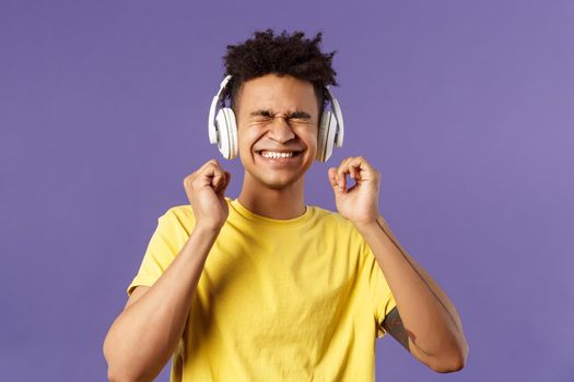 Portrait of pleased, excited young man enjoying nice quality awesome beats in headphones, close eyes and smiling rejoicing, dancing over cool new song, listen music over purple background.