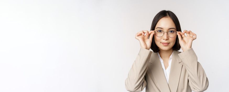 Smiling asian businesswoman trying new glasses, wearing eyewear, standing in suit over white background. Copy space