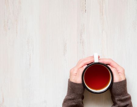 Female hands holding a mug of hot black tea. Cold winter, warm clothes, sweater. Top view. White aged wooden background