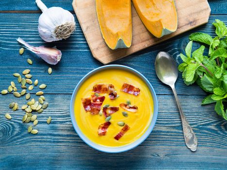 Creamy soup of pumpkin with bacon and seeds on dark blue wooden background, garlic, mint, slices of pumpkin. Top view.