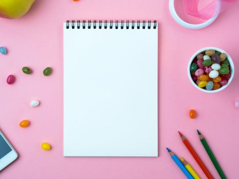 Open notebook with a clean white sheet, sweets, mobile phone, crayon, decorations on a pink bright table. Girl's workplace for creativity, plans and dreams