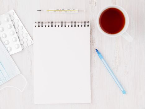 open notebook with blank white clean page to write the treatment plan of the illnedd. Pills, pen, paper, thermometer on white wood background, top view.