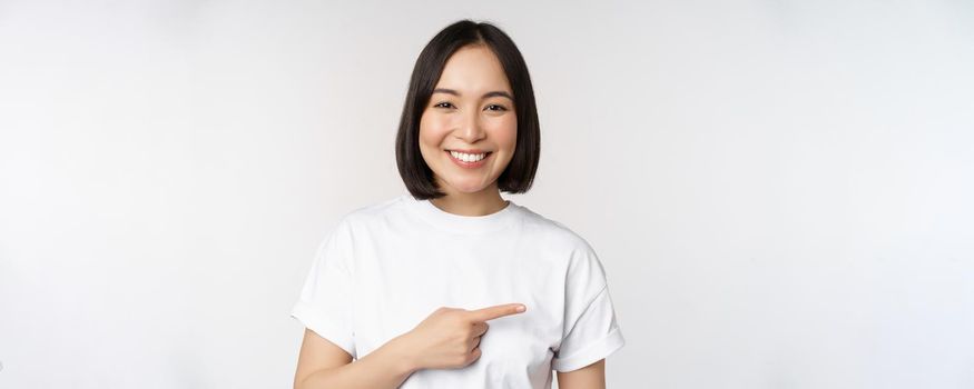 Happy beautiful asian girl showing advertisement on empty copy space, pointing finger right and smiling, demonstrating banner or company logo, white background.