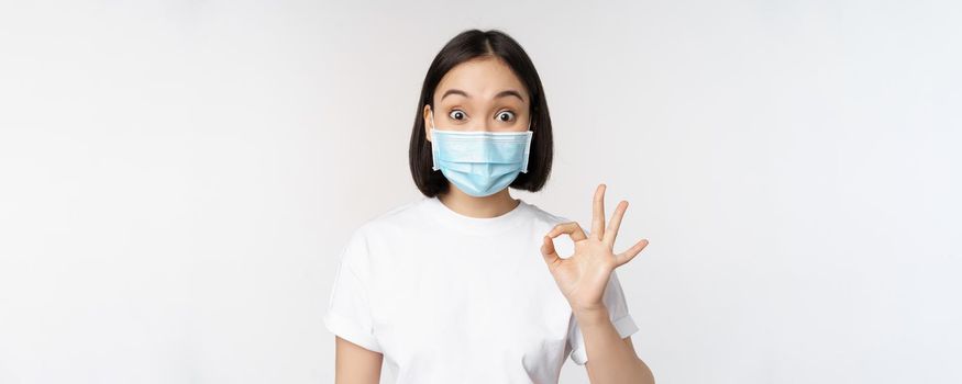 Covid-19, healthcare and medical concept. Impressed asian woman in medical mask, looking amazed and showing ok, okay sign, standing over white background.