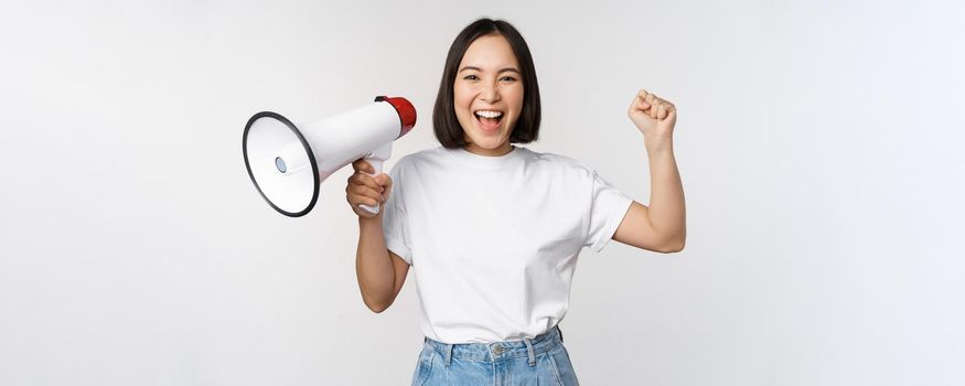 Happy asian woman shouting at megaphone, making announcement, advertising something, standing over white background.