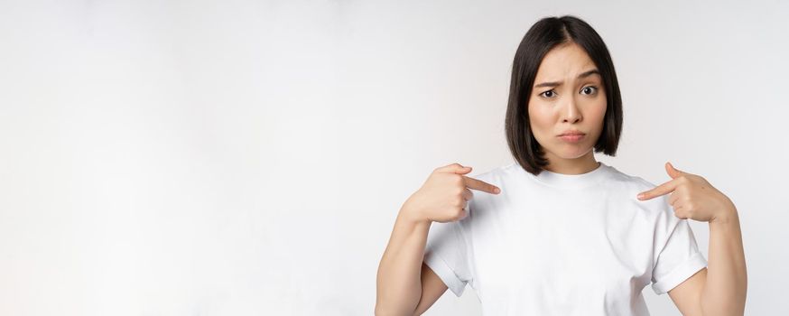 Young asian woman pointing at herself with disbelief, being chosen, surprised by her candidature, standing over white background.