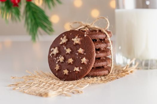 a stack of round chocolate Christmas cookies with white sugar stars and salt tied with a rope, a glass of milk , stands on a white table, under a spruce branch, shining garlands in the background