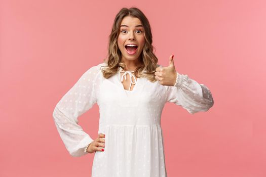Great idea. Upbeat, enthusiastic blond caucasian girl in white spring dress, hear good plan, show thumbs-up and nod in approval, agree and recommend, satisfied with choice, stand pink background.
