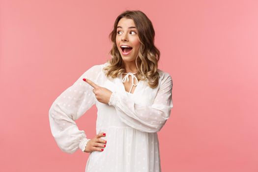 Excited and amazed blond caucasian girl in kawaii dress, open mouth gasping in awe, pointing and looking left with excitement and astonishement, see great beauty product, pink background.