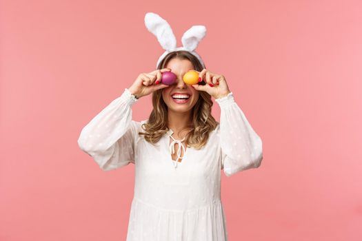 Holidays, spring and party concept. Portrait of lovely, tender smiling woman in rabbit ears and white dress celebrating Easter day, holding painted eggs on eyes and grinning, pink background.