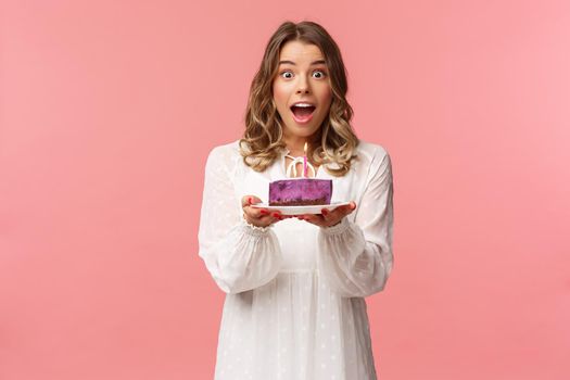 Holidays, spring and party concept. Happy cheerful good-looking blond woman celebrating birthday, holding piece cake with lit candle, making wish, look amused standing pink background.