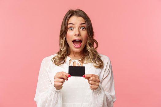 Close-up portrait of emotive, excited and thrilled good-looking blond girl showing credit card and feeling very happy, telling about discounts, special offers, bank service, pink background.