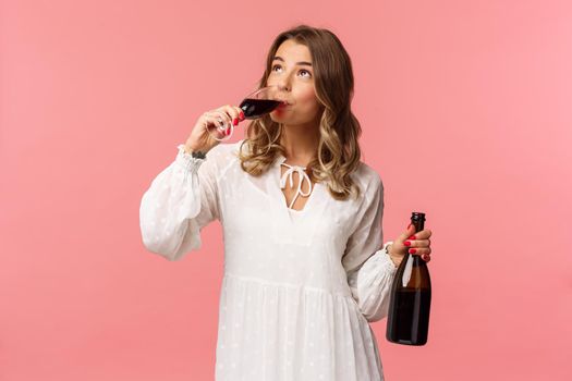 Holidays, spring and party concept. Portrait of young elegant happy blond woman, wear white trendy dress, drinking wine from glass looking up pleased, tasting good drink, hold bottle.