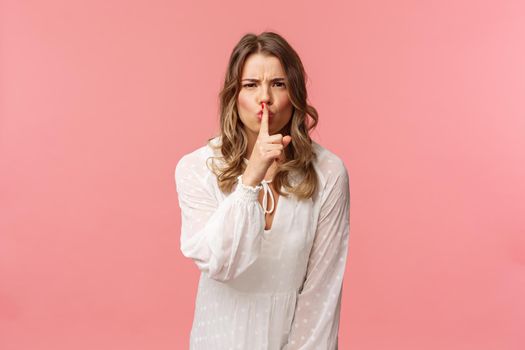 Beauty, fashion and women concept. Angry serious-looking blond girl in white dress, shushing at someone making noise, press finger to lips scolding bad behaviour, say be silent, pink background.