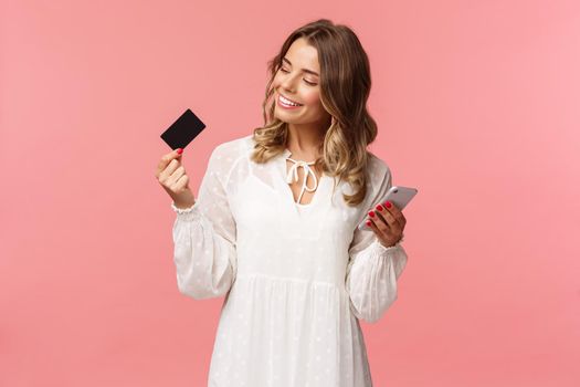 Portrait of happy good-looking young blond woman in white dress, looking pleased and satisfied at credit card, holding smartphone, recommend buy online, internet shopping concept.