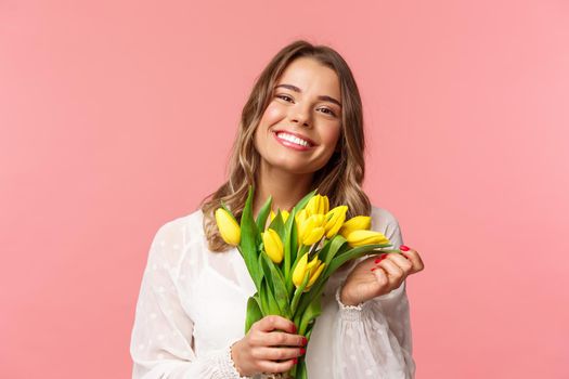 Spring, happiness and celebration concept. Close-up of lovely happy young blond girl in white dress, daydreaming after perfect date, holding yellow tulips and smiling at camera, pink background.