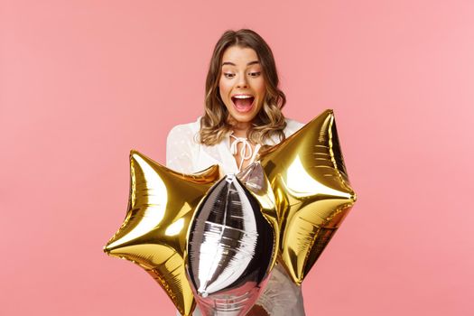 Holidays, celebration and women concept. Portrait of happy lovely young woman in white dress, gasping from amazement and joy, holding birthday star-shaped balloons, pink background.