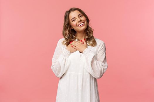 Beauty, fashion and women concept. Portrait of feminine blond young woman in white cute dress, hold arms pressed to heart and sighing from admiration and delight, feel thankful, pink background.