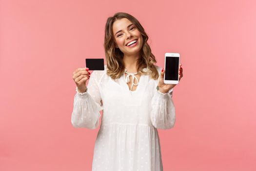 Portrait of tender feminine young blond girl in white dress, tilt head and smiling pleased, advice download application, holding mobile phone, credit card, show smartphone screen, pink background.