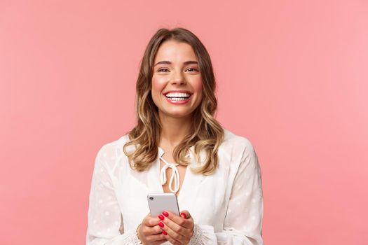 Close-up portrait of charming, lovely blond girl using mobile phone, texting friend, order in online store using smartphone application, laughing and smiling pleased, stand pink background.