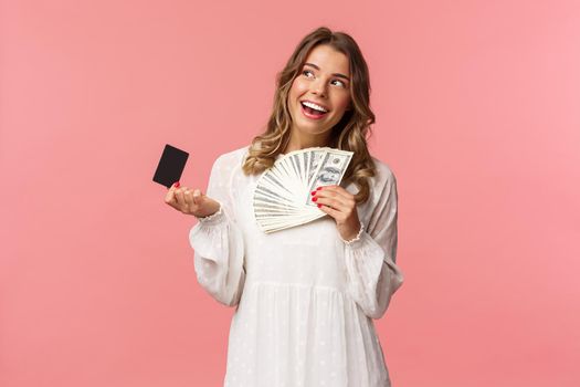 Portrait of pleased attractive blond girl in white dress, daydreaming about what buy, shopping with girlfriends, holding money and credit card, use cash, standing pink background.