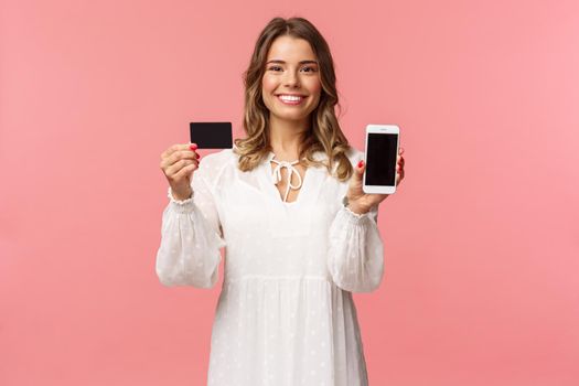Portrait of satisfied cute blond girl in white dress, showing you her credit card and mobile phone application, online shopping app or bank website, smiling camera, standing pink background.