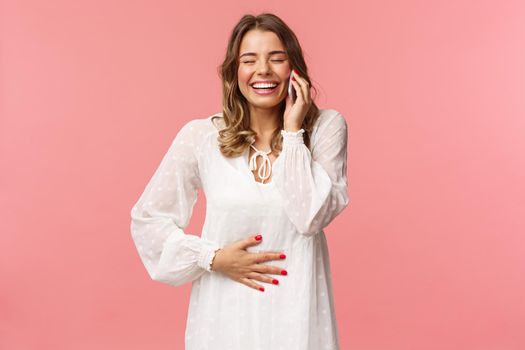 Portrait of joyful happy bright young girl in white dress, having fun, speaking to friend, calling person mobile phone, laughing and touching belly, close eyes smiling, hold smartphone near ear.