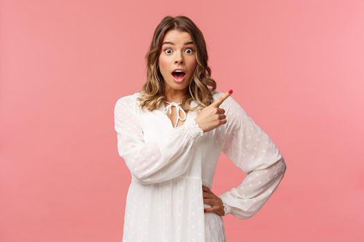 Surprised beautiful blond girl pointing upper right corner, gasping amused and staring at camera with fascinated expression, open mouth in awe to ask question about promo, pink background.