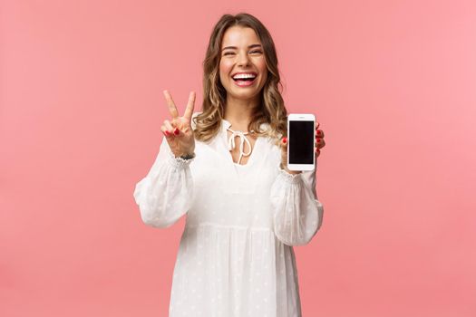 Portrait of kawaii optimistic and happy young girl in white dress, show mobile phone display and peace sign, laughing feeling cheerful and glad to share awesome app, useful link, pink background.