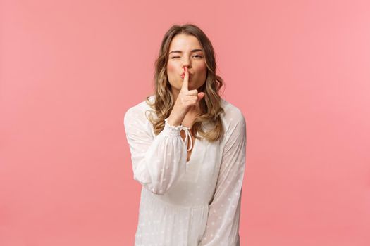 Beauty, fashion and women concept. Portrait of feminine and flirty caucasian woman hiding secret, asking keep silent, shushing with sensual coquettish smile, wear white dress, pink background.