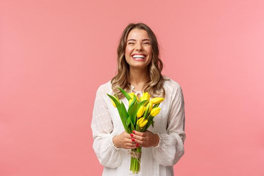 Holidays, beauty and spring concept. Portrait of happy excited charming blond girl receive flowers, buying yellow tulips herself, smiling and laughing joyfully, stand pink background.