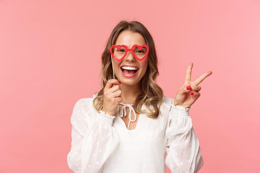 Spring, happiness and celebration concept. Close-up portrait of upbeat blond girl at party, wear white dress holding heart-shaped glasses mask over eyes and make peace sign cheerful.
