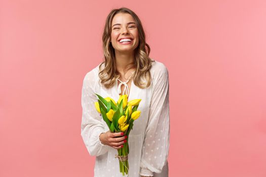 Holidays, beauty and spring concept. Carefree happy attractive blond girl in white dress holding yellow tulips and laughing with pleased expression, standing pink background, have romantic date.