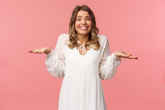 Beauty, fashion and women concept. Sorry dont know, cant say. Cheerful cute silly blond girl 20s, wear white dress, shrugging with hands sideways make awkward smile, apologizing, pink background.