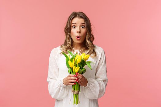 Holidays, beauty and spring concept. Portrait of surprised and amazed blond girl in white dress, holding yellow tulips, receive flowers being amused and happy, standing pink background.