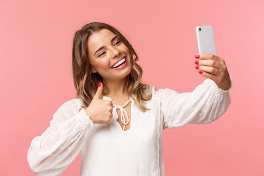 Close-up portrait of stylish beautiful beauty blogger record video on smartphone, taking selfie, showing thumbs-up at mobile phone camera, smiling pleased, recommend place to followers.