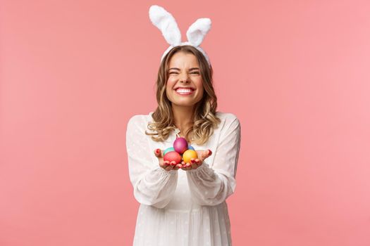 Holidays, spring and party concept. Portrait of excited charming blond woman with lovely pleased smile, holding painted eggs, wear Easter day outfit, rabbit ears and white dress, pink background.