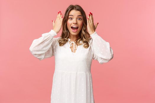 Surprised happy, excited b-day girl in white dress open her eyes to see incredible awesome gift from friends at party, standing over pink background look astonished and cheerful.