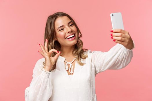 Close-up of satisfied good-looking blond girl in white dress, taking selfie, record mobile phone video, show okay satisfactory sign with pleased nod, smiling agree or recommend, pink background.