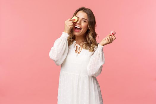 Holidays, spring and party concept. Portrait of carefree, tender and feminine girl in white dress, holding macarons dessert over eye, feeling cheerful, eating sweets, tasty desserts, pink background.