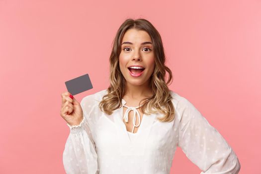 Close-up portrait of excited pretty young blond woman in white dress, holding credit card, showing people how she invests, look camera amused, smiling recommend shopping app, pink background.