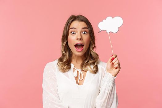 Spring, happiness and celebration concept. Close-up portrait of surprised, excited blond girl looking amazed, holding cloud comment stick near head and gasping astonished, have idea, pink background.