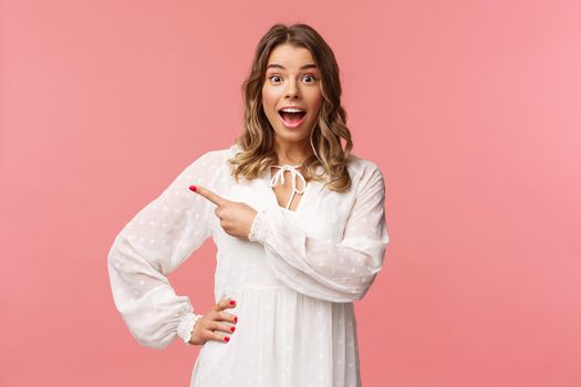 Excited and amused, intrigued blond european girl in white cute spring dress, open mouth wondered and amazed of hearing awesome news, pointing finger left at something cool, pink background.